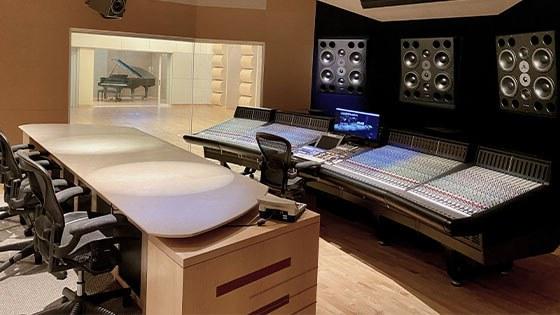 A conference table faces a wall with mixing boards and speakers; a recording area with a grand piano is in the background.